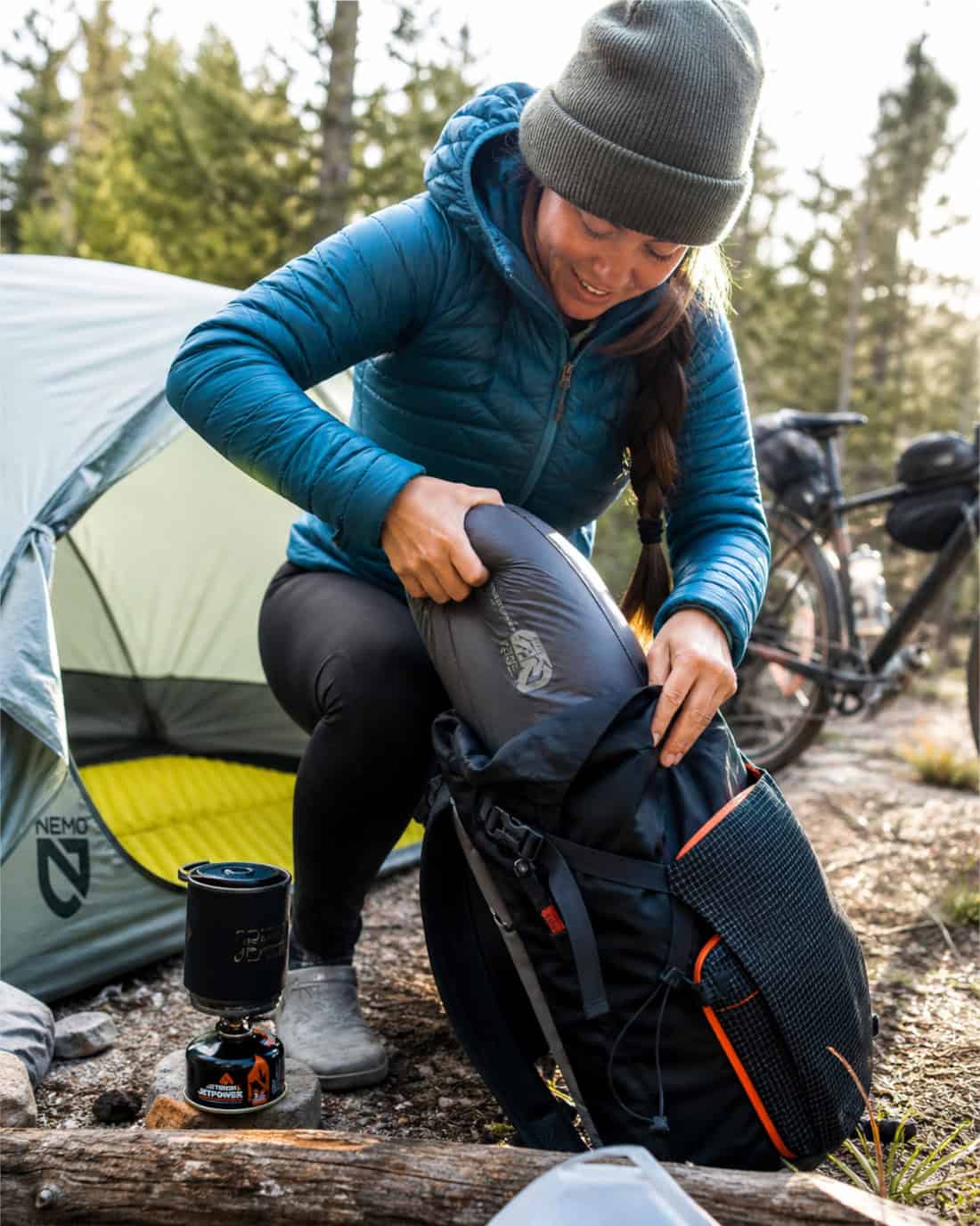 A camper stuffs a compact sleeping bag into a backpack beside a tent and bikepacking setup, with bright morning light and an energetic ready-to-go camping mood